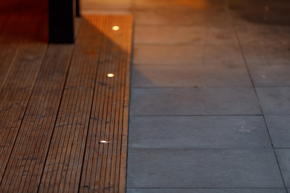 Drewniany taras z subtelnym oświetleniem punktowym, graniczący z szarą, betonową nawierzchnią. Ciepłe światło lamp kontrastuje z chłodnym odcieniem betonu.