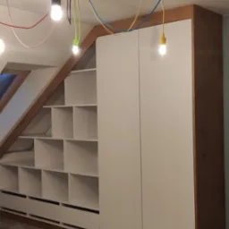 Szafa wnękowa z półkami i szufladami na poddaszu, białe fronty, drewniane wykończenie, oświetlenie z kolorowymi kablami i żarówkami.