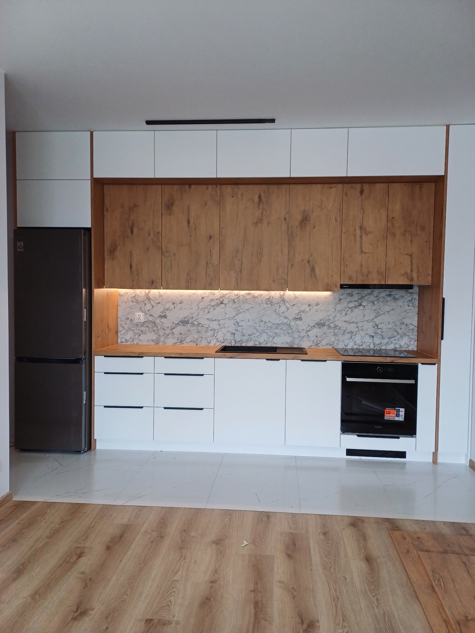 Nowoczesna kuchnia: białe szafki dolne z czarnymi uchwytami, drewnopodobne szafki górne, marmurowy backsplash, czarny sprzęt AGD i podświetlenie LED.