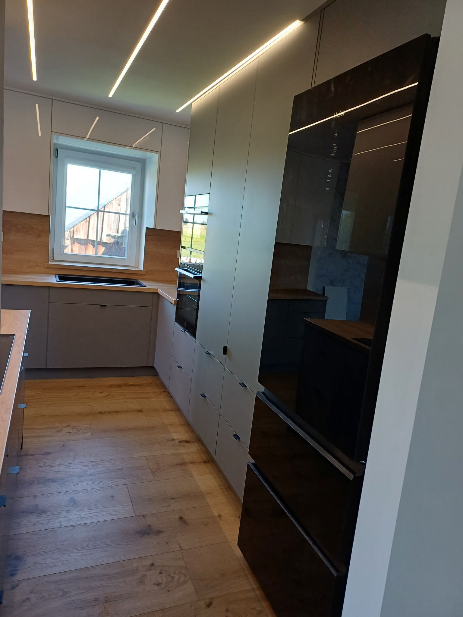 Nowoczesna kuchnia z szarymi szafkami i drewnianym blatem, oświetlona ledami. Widoczna duża, czarna lodówka z połyskiem i drewniana podłoga.