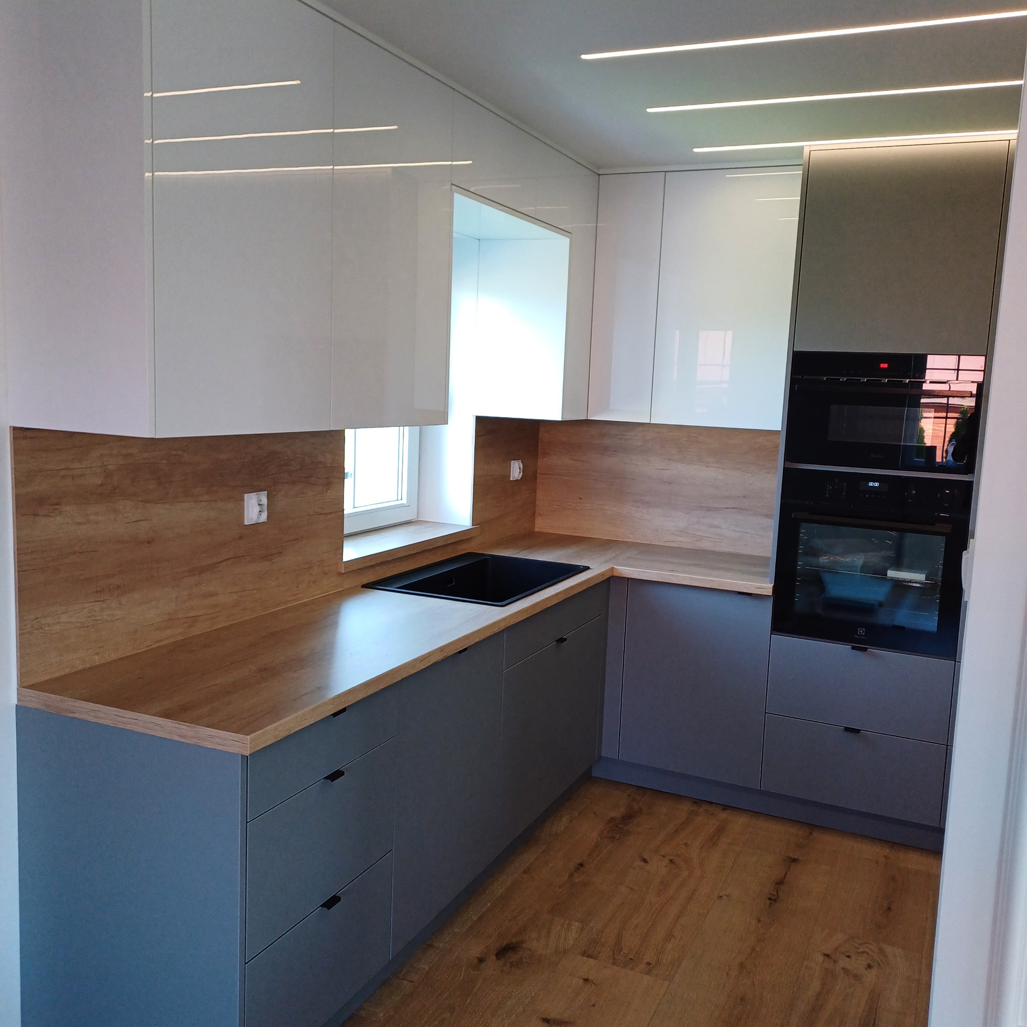 Nowoczesna kuchnia na wymiar: szare szafki dolne z drewnianym blatem, białe szafki górne z oświetleniem LED, zabudowany piekarnik i mikrofalówka.