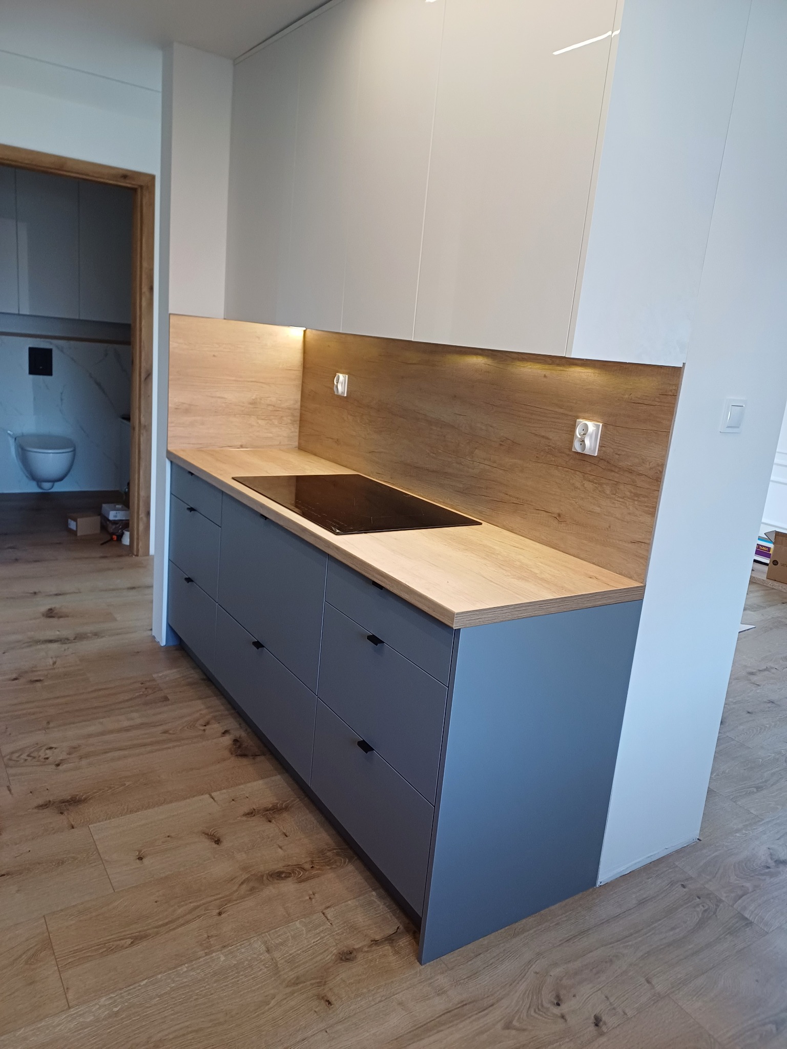 Szare szafki kuchenne z drewnianym blatem i panelem, oświetlenie LED pod szafkami, widok na łazienkę w tle, drewniana podłoga.