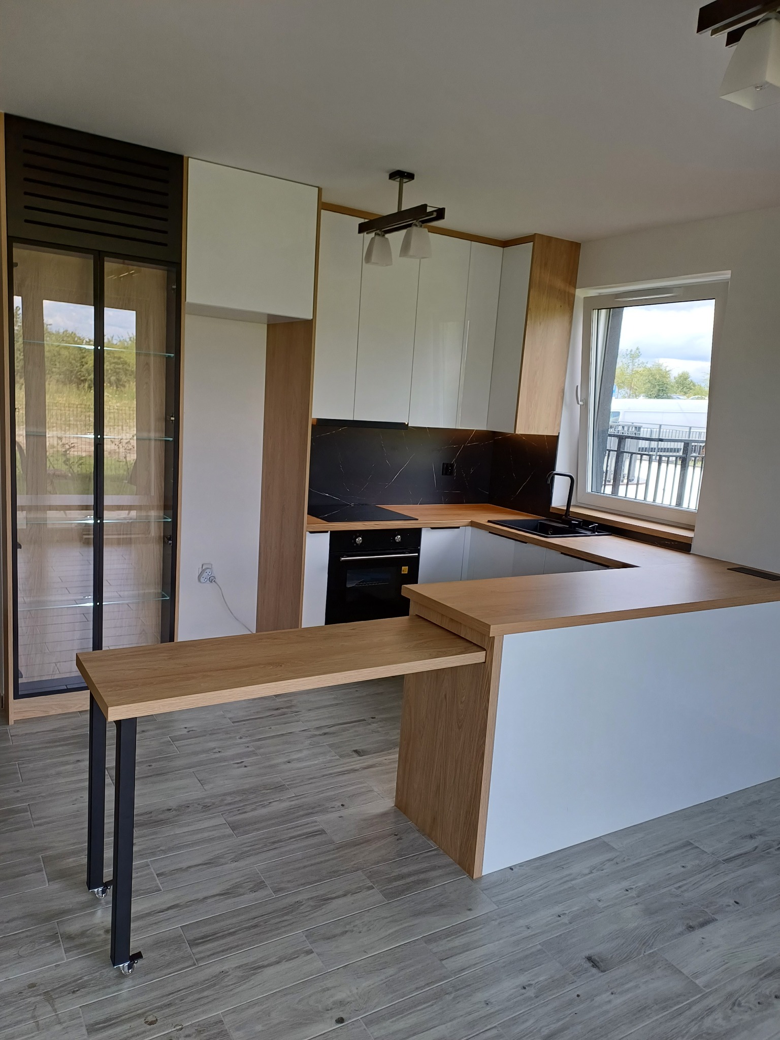 Nowoczesna kuchnia z wysuwanym blatem, białe fronty szafek, drewniane akcenty i czarny panel nad blatem. Widoczna przeszklona witryna i okno.