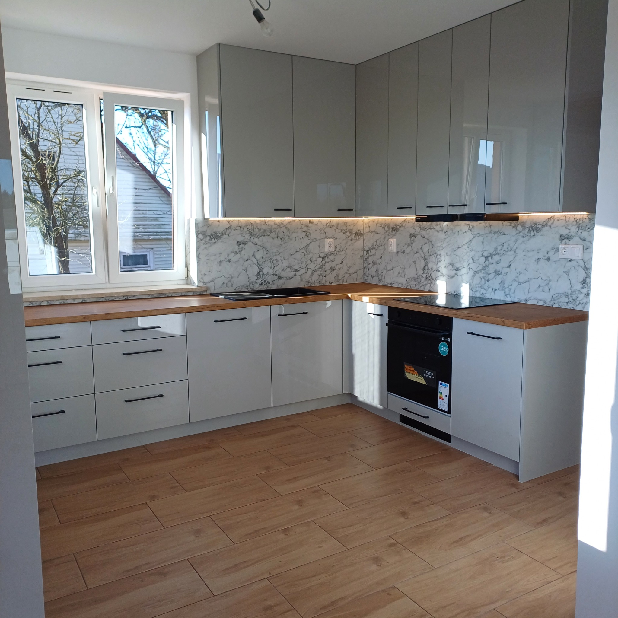 Jasnoszara kuchnia z drewnianym blatem i marmurowym panelem nad blatem, oświetlona listwą LED. Widok na okno z drzewem i domem w tle. Nowoczesny design.