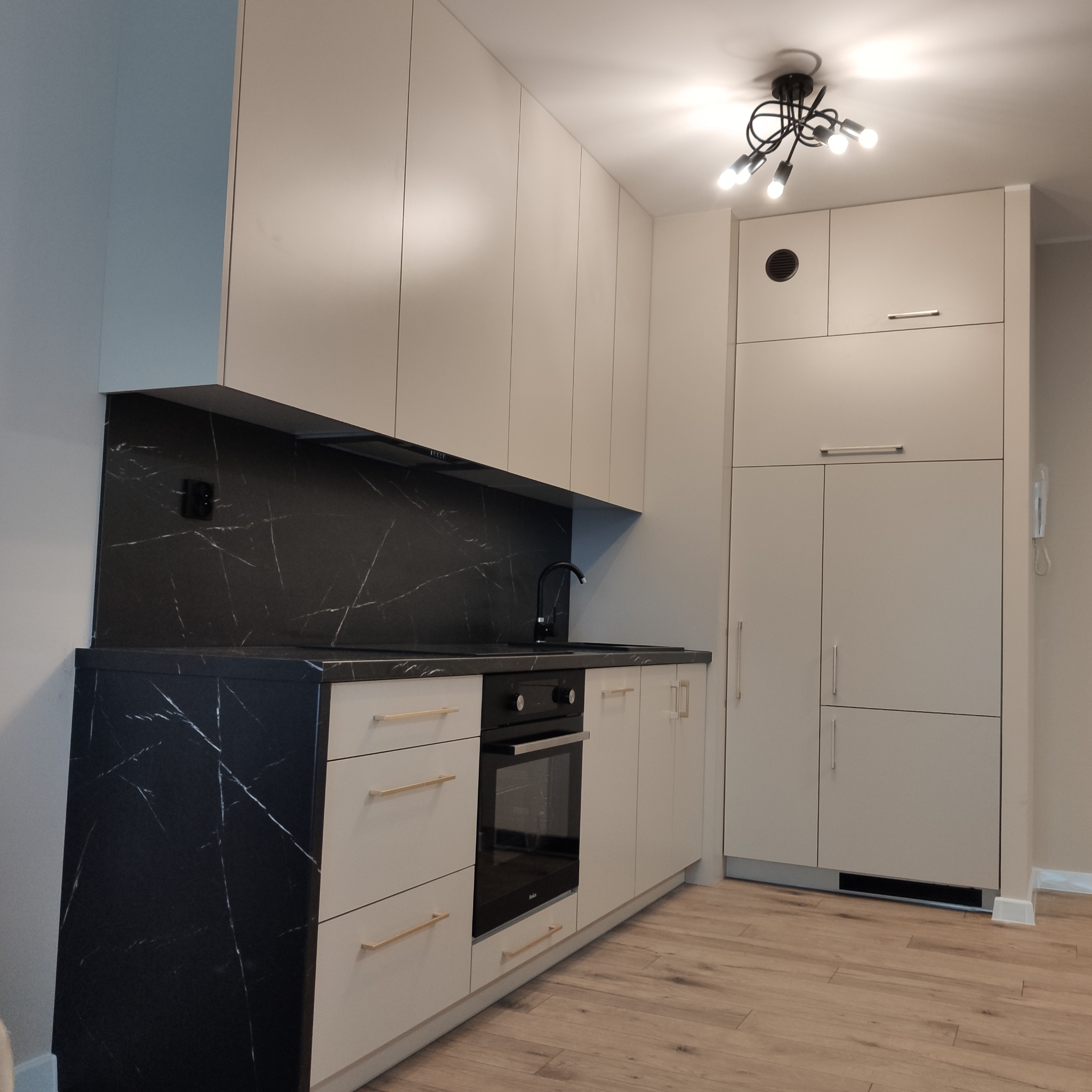 Kremowa kuchnia z blatem imitującym czarny marmur, złote uchwyty, czarna armatura i oświetlenie sufitowe. Fronty gładkie, bez zdobień.