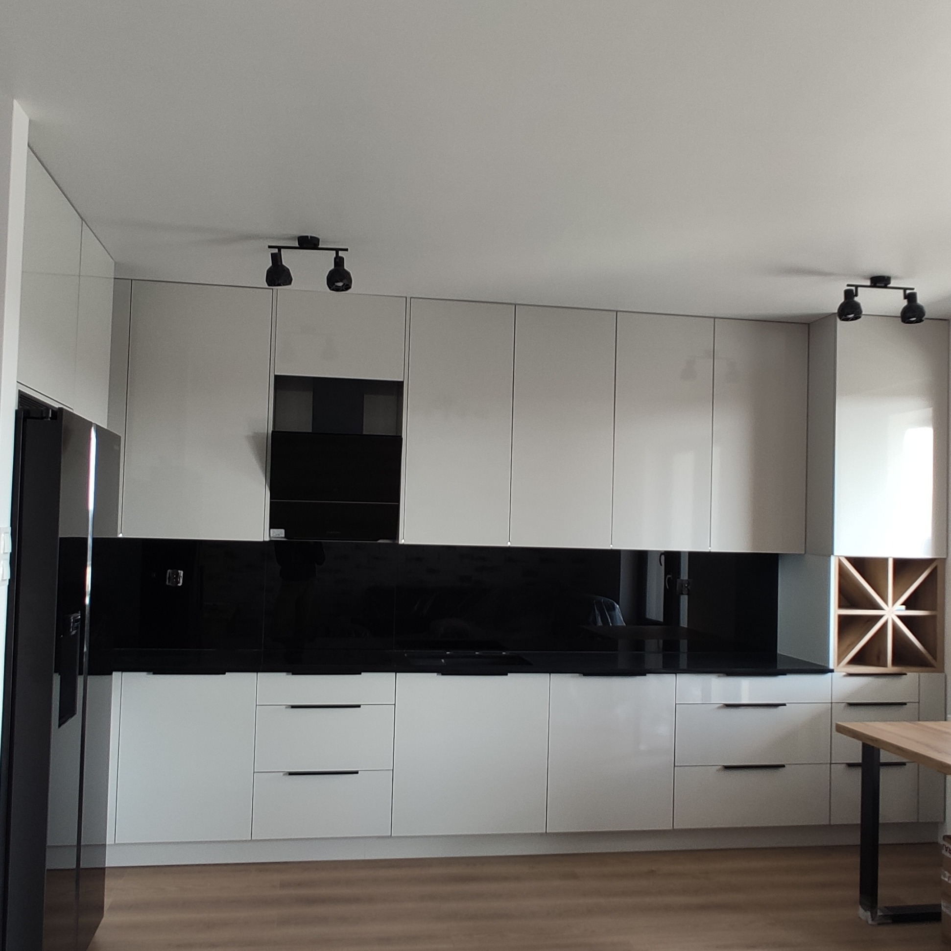 Biała kuchnia z czarnym panelem szklanym nad blatem i drewnianym regałem na wino. Minimalistyczny design, oświetlenie punktowe.