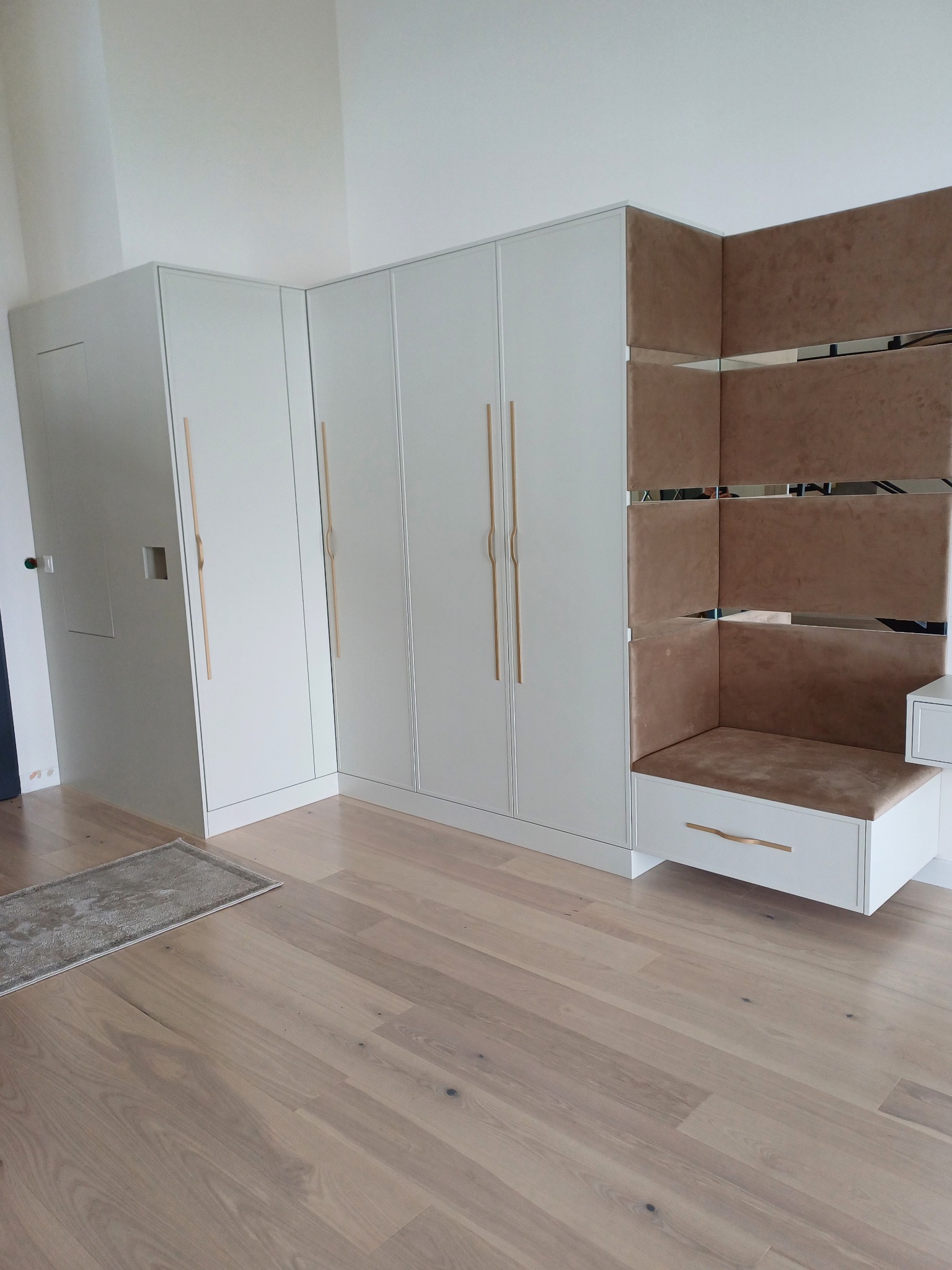 Biała szafa wnękowa z tapicerowanym siedziskiem i lustrzanymi wstawkami, złote uchwyty, jasna podłoga. Minimalistyczny design, nowoczesne wykończenie.