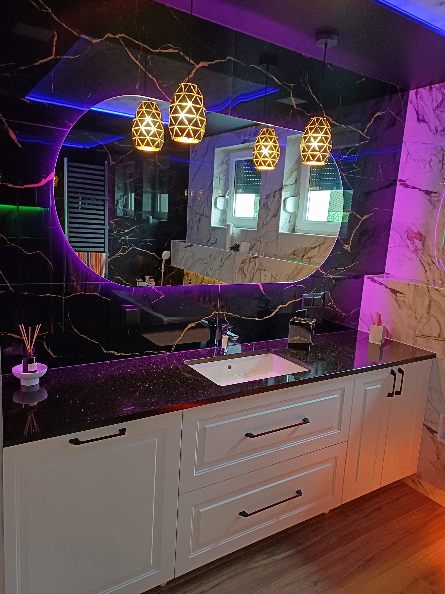 Nowoczesna łazienka z białymi szafkami pod umywalką, czarnym blatem i okrągłym lustrem podświetlonym fioletowym światłem, wiszące lampy nad lustrem.