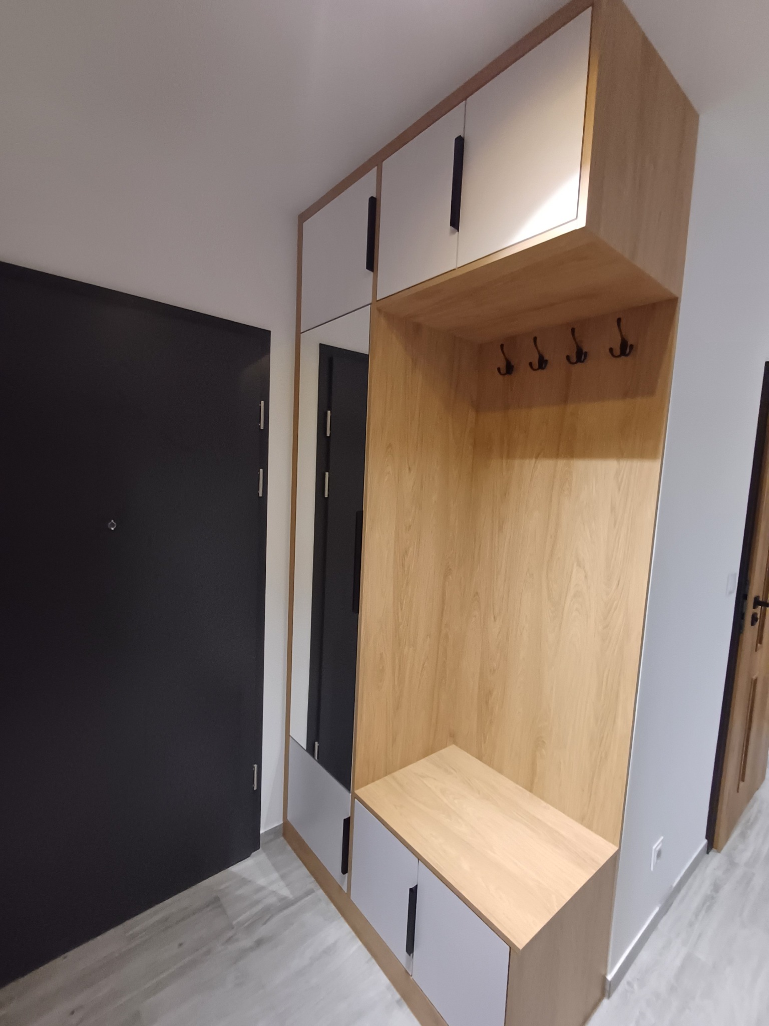 Funkcjonalna szafa wnękowa z lustrem i wieszakami, wykończona w drewnie i bieli, idealna do przedpokoju. Zwraca uwagę minimalistyczny design i praktyczne rozwiązania.