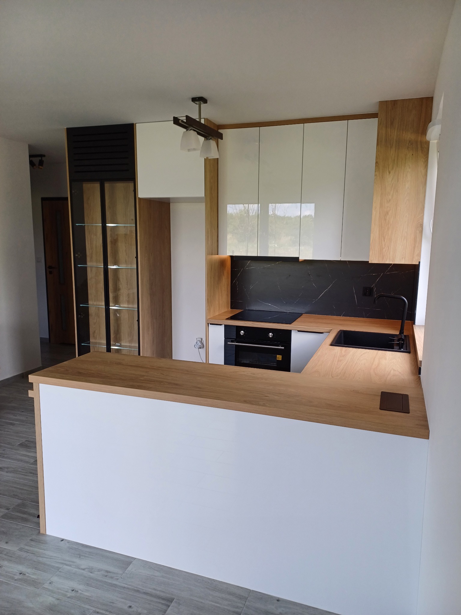 Nowoczesna kuchnia z białymi szafkami, drewnianym blatem i czarnym panelem. Widoczna wyspa kuchenna i przeszklona witryna. Oświetlenie podszafkowe.