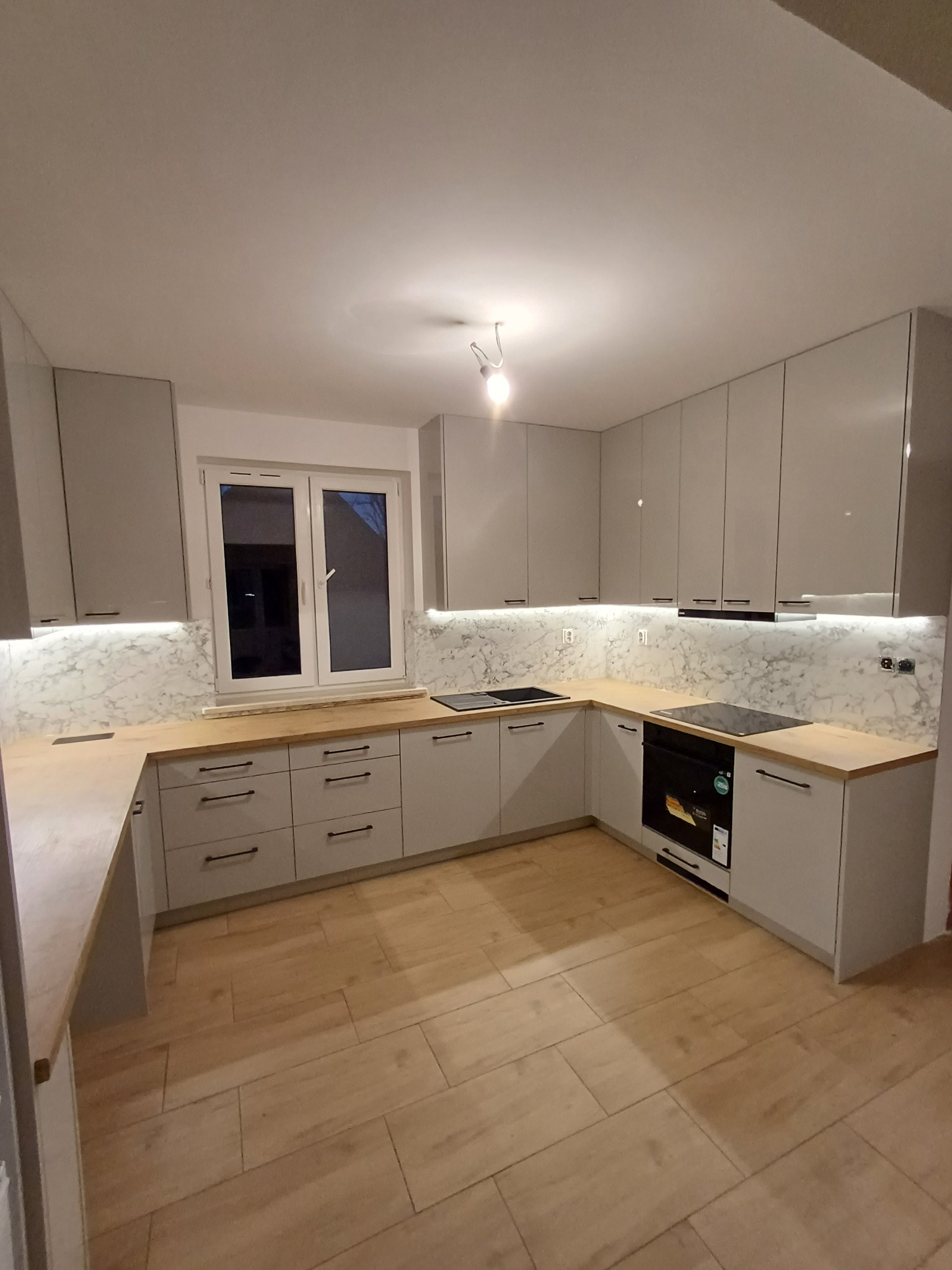 Jasna kuchnia z drewnianym blatem i marmurowym backsplashiem. Górne szafki bez uchwytów, oświetlenie LED pod szafkami. Nowoczesny design, drewniana podłoga.