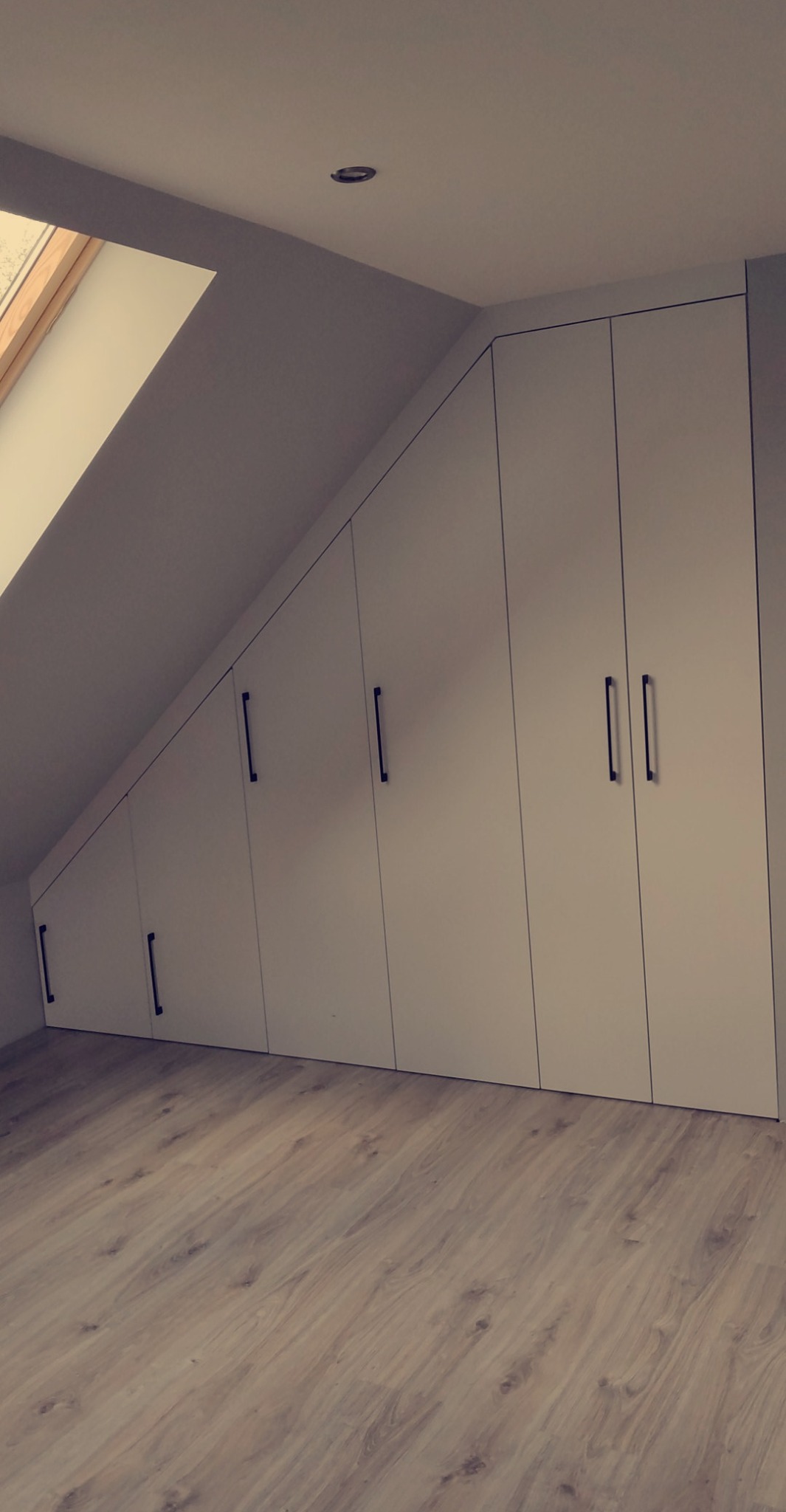 Białe szafy wnękowe z czarnymi uchwytami, dopasowane do skosu poddasza, na jasnej podłodze. Widok z dołu, okno dachowe w tle.