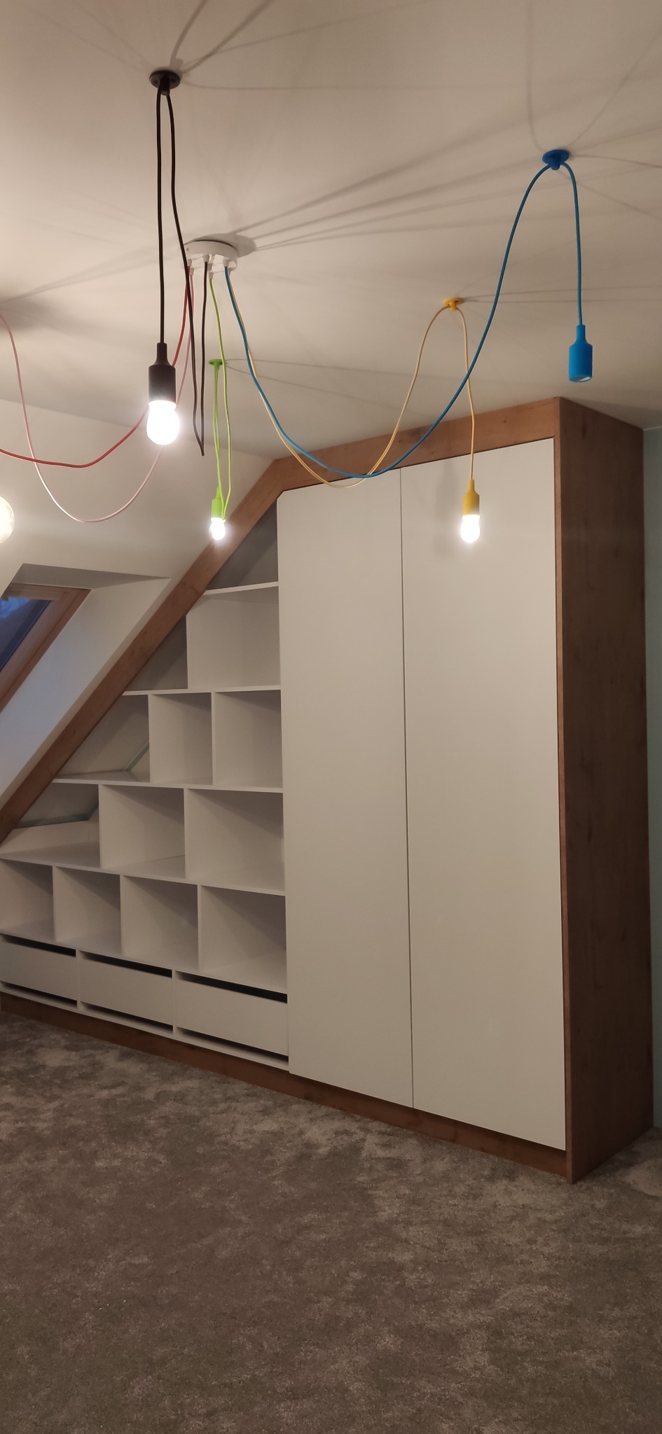 Szafa wnękowa z półkami i szufladami na poddaszu, białe fronty, drewniane wykończenie, oświetlenie z kolorowymi kablami i żarówkami.