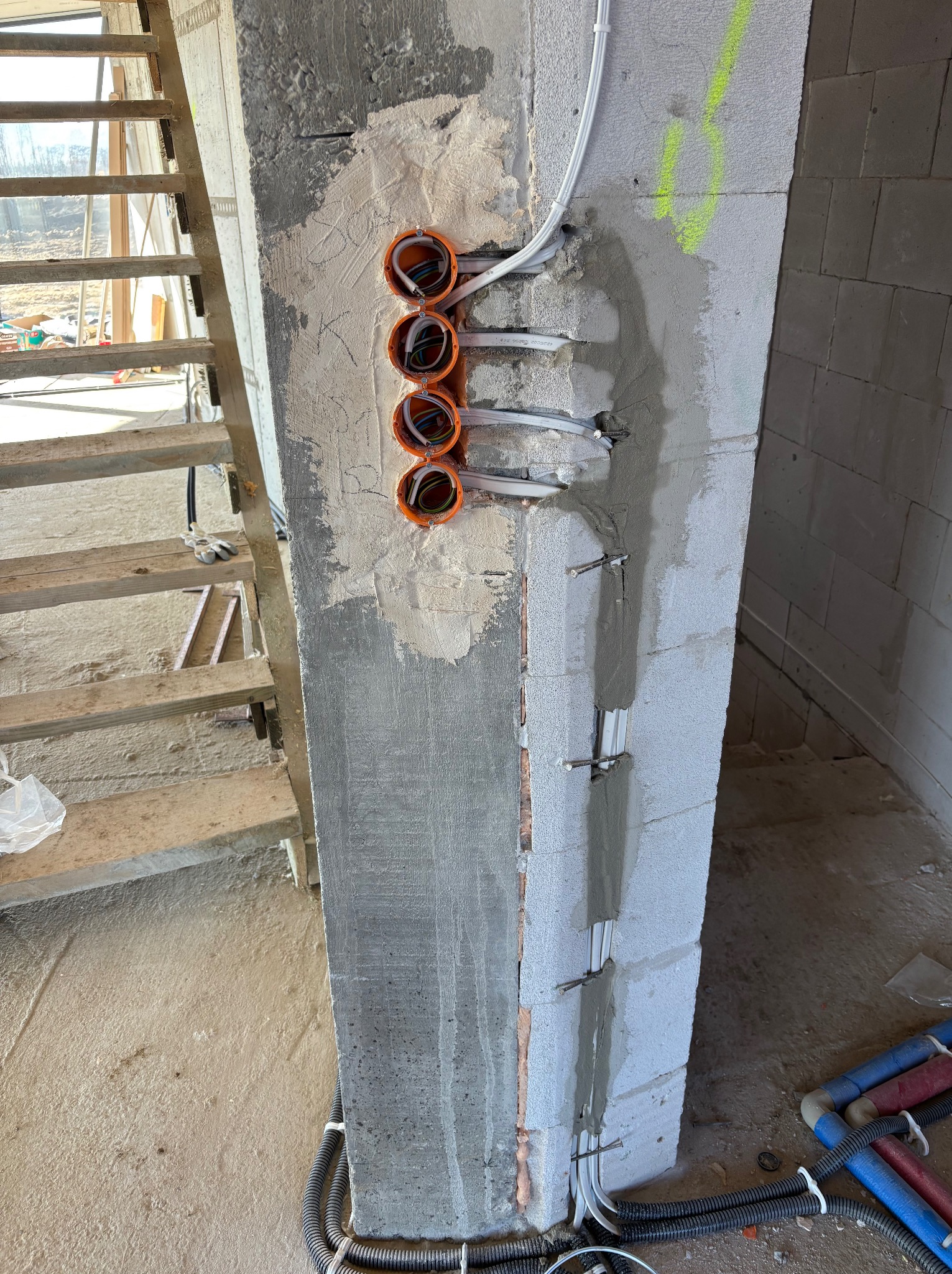 Instalacja elektryczna w trakcie montażu: pomarańczowe puszki podtynkowe z przewodami w ścianie z betonu komórkowego, kable zabezpieczone klamrami. Widoczne schody.