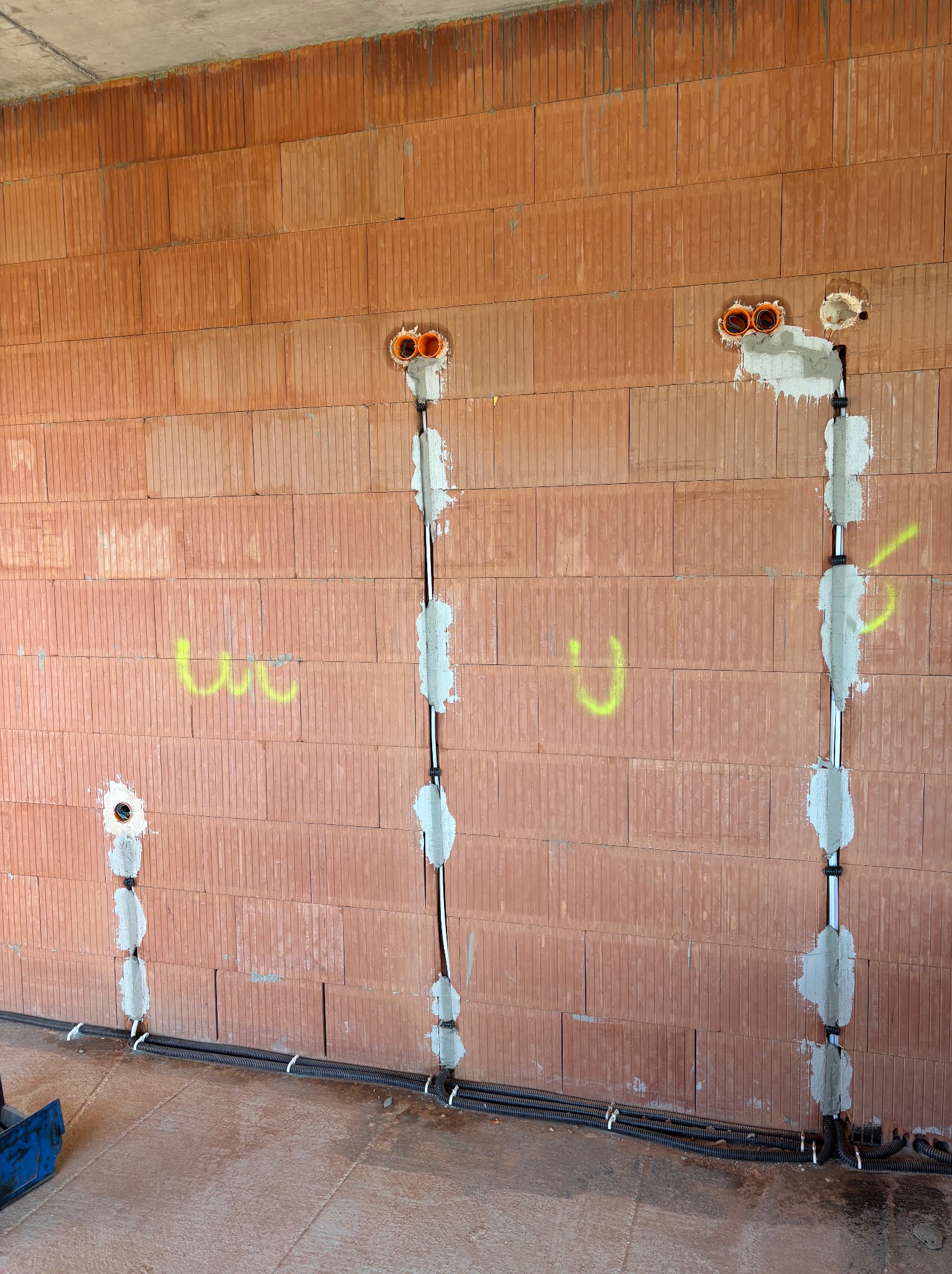 Instalacja elektryczna w trakcie montażu w nowym budynku z czerwonej cegły. Widoczne kable w peszlach, puszki pod gniazdka i oznaczenia na ścianie.