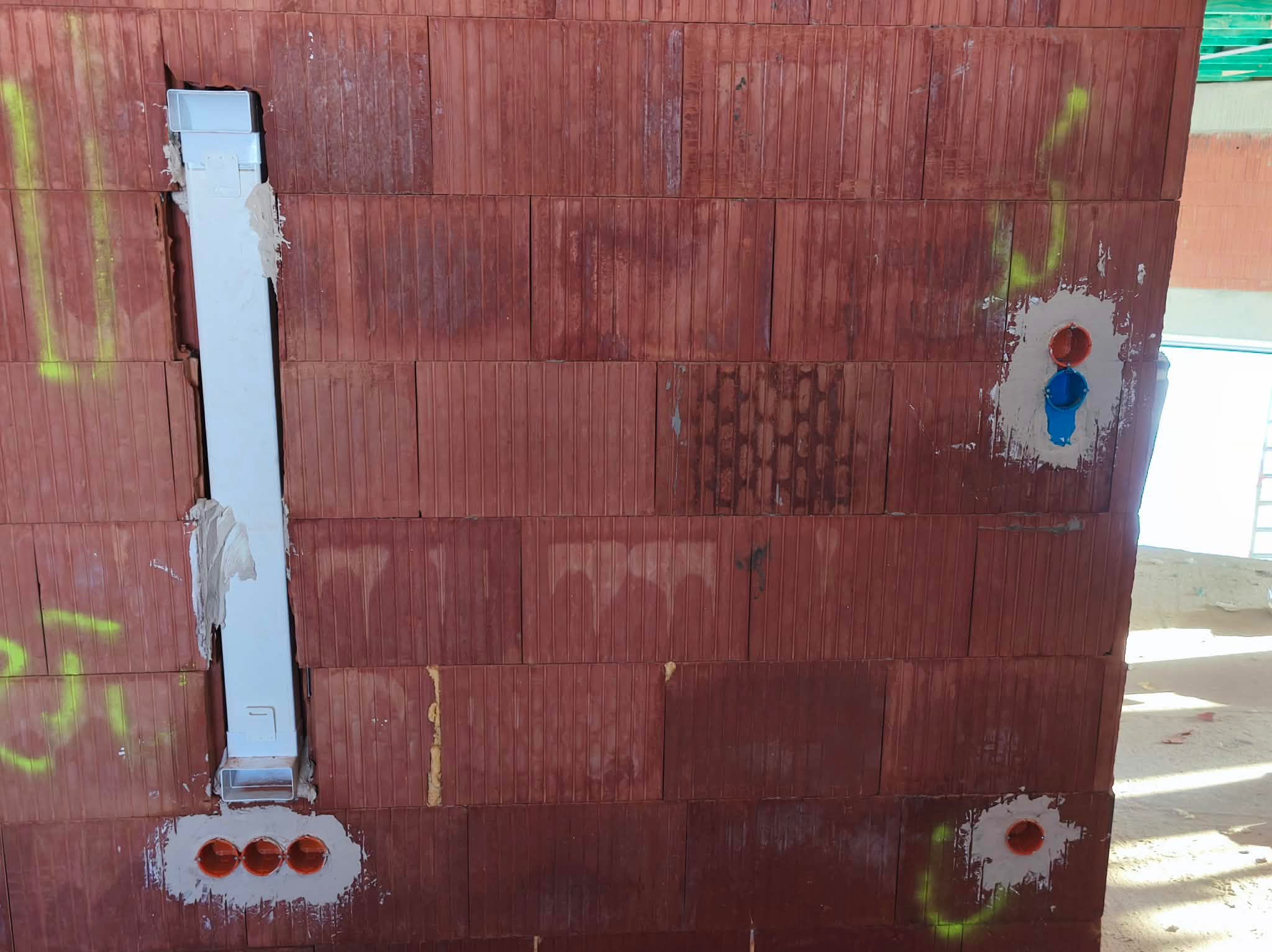 Czerwona ściana z pustaków z zamontowaną, pionową, białą rurą instalacyjną i otworami na puszki elektryczne. Widoczne ślady zaprawy i żółte oznaczenia.