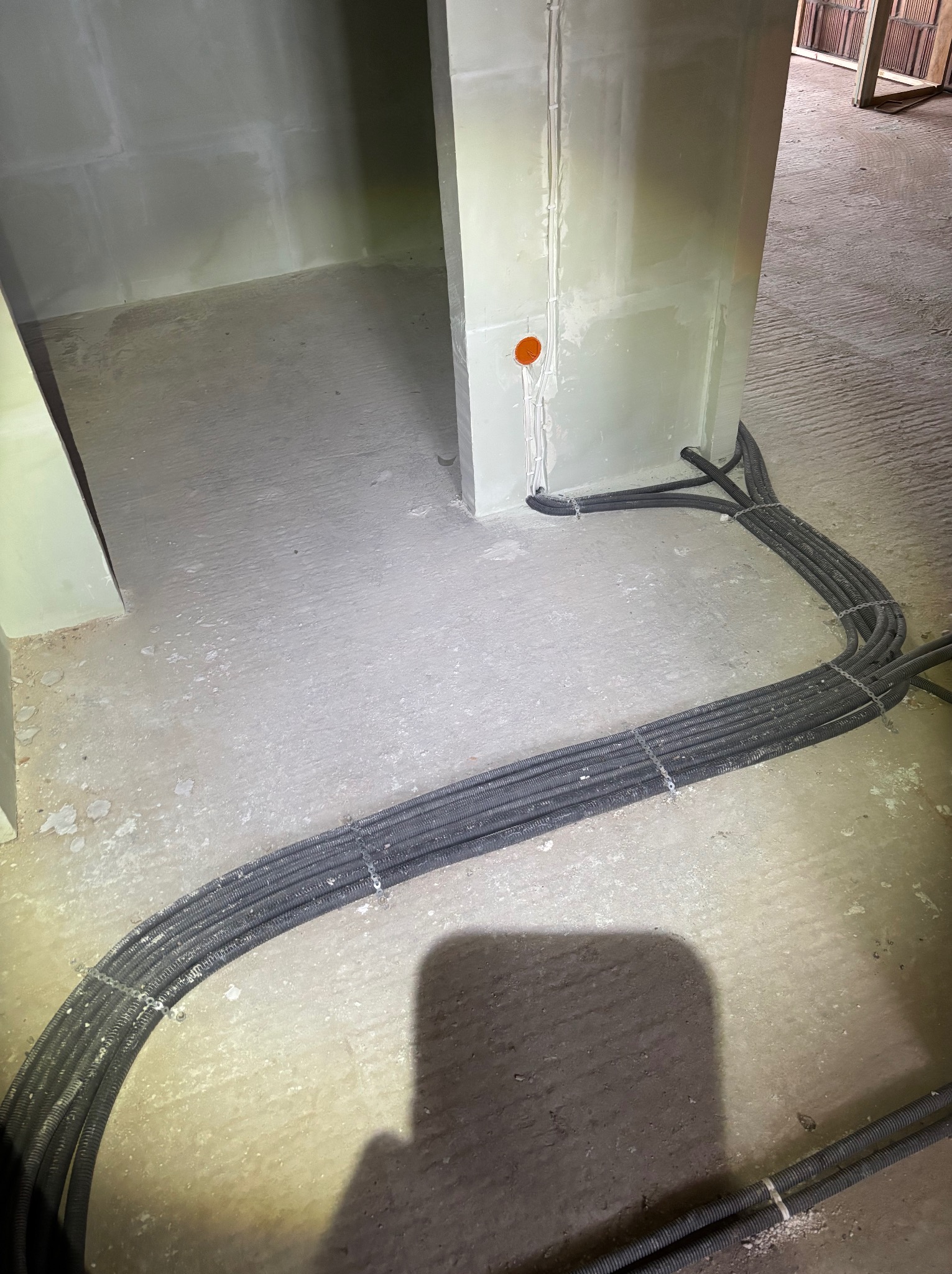 Kable elektryczne w peszlach ułożone na podłodze w surowym wnętrzu, prowadzące do ściany z zamontowaną puszką elektryczną. Widoczne mocowania kabli.