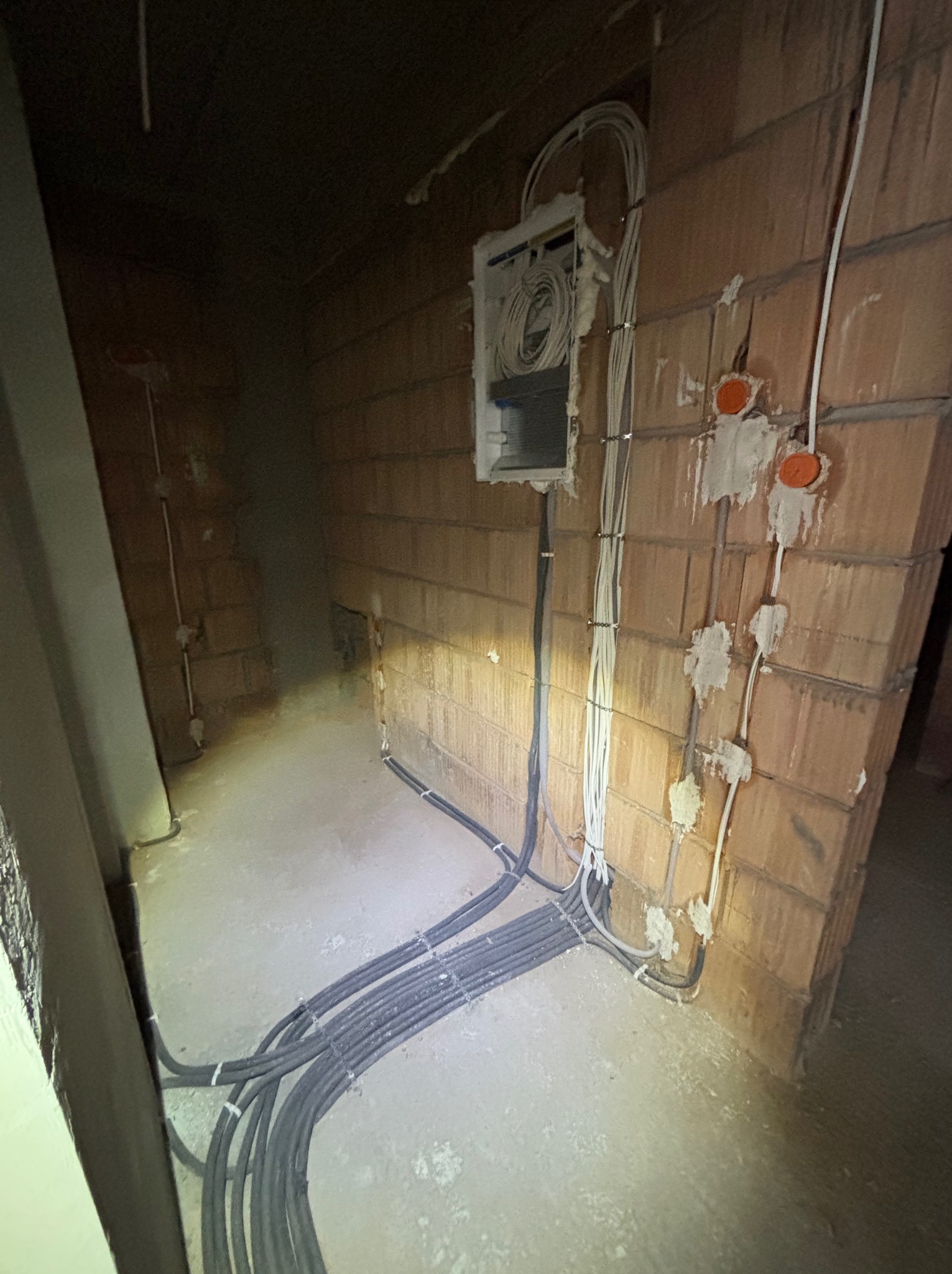 Surowa instalacja elektryczna w budynku z cegły: kable w peszlach prowadzone po podłodze i ścianach, puszki instalacyjne zamontowane, widoczne przewody. Etap prac przed wykończeniem.