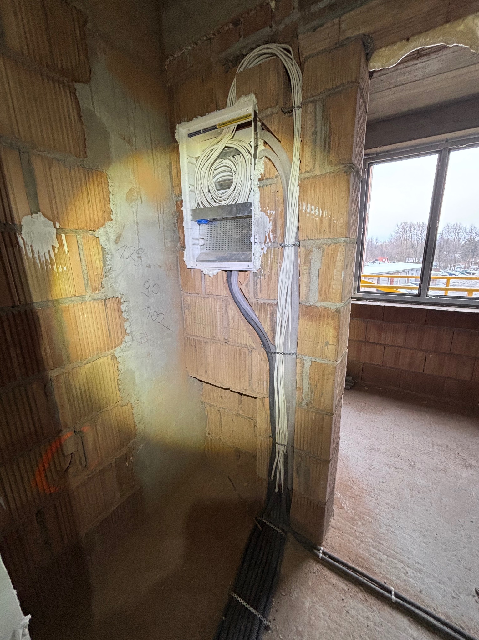 Rozdzielnia elektryczna z kablami w surowym wnętrzu budynku z cegły, widok na okno i krajobraz za nim. Instalacja w trakcie realizacji.
