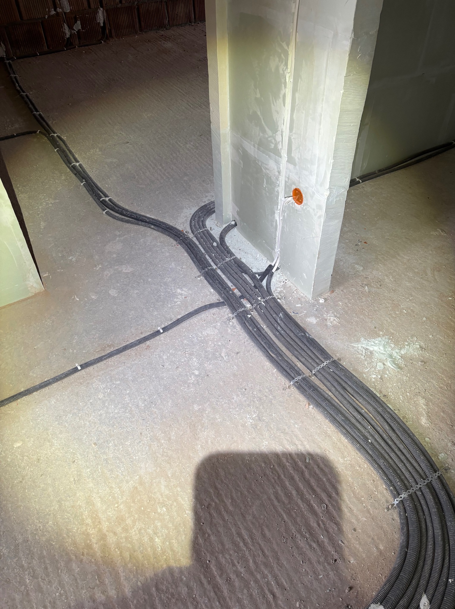 Kable elektryczne w peszlach ułożone na surowym betonie wzdłuż ściany z gniazdkiem. Widoczne mocowania kabli. Etap prac instalacyjnych w budynku.