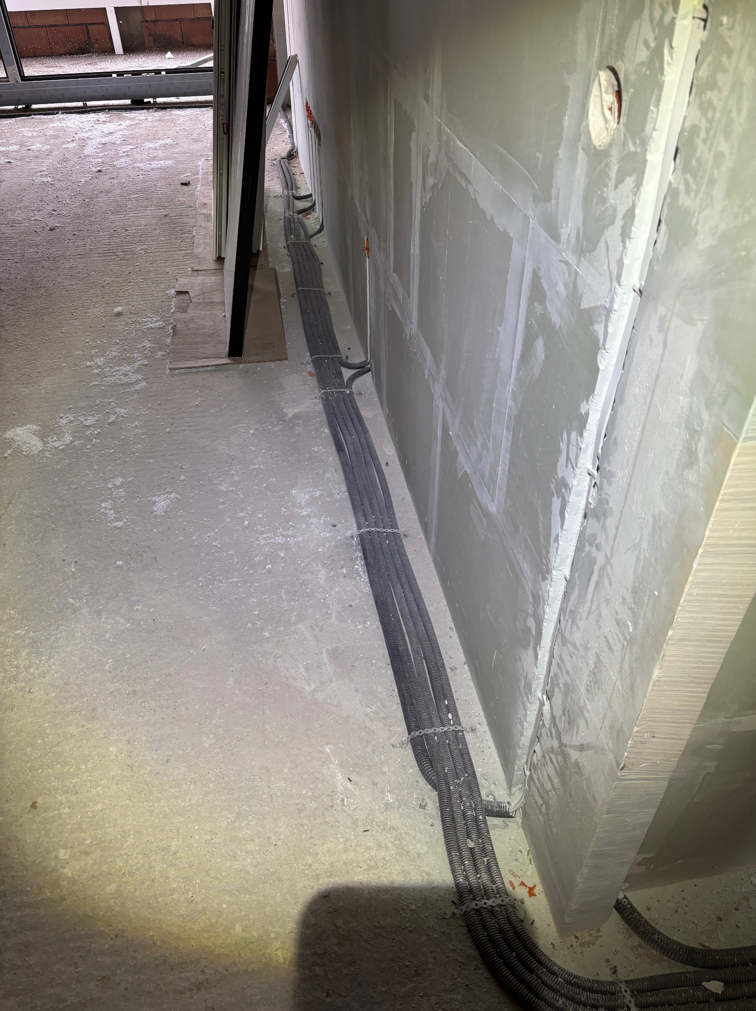 Szare kable elektryczne w peszlu, ułożone wzdłuż ściany w surowym wnętrzu. Widoczne puszki instalacyjne w ścianie i fragment podłogi z resztkami materiałów budowlanych.