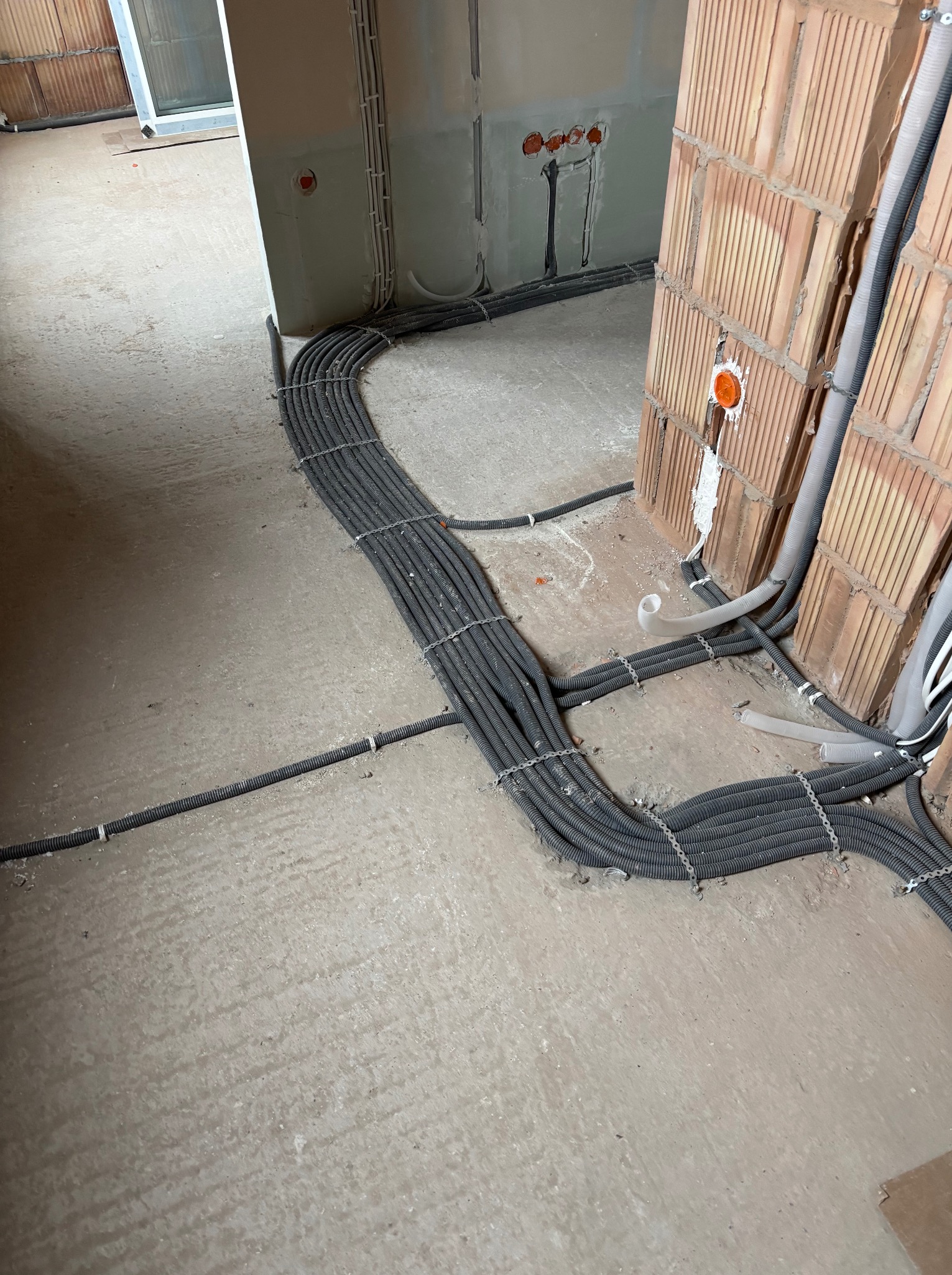 Szare rury instalacji elektrycznej ułożone na podłodze w surowym stanie, prowadzone wzdłuż ściany z cegły dziurawki i ściany otynkowanej. Widoczne puszki instalacyjne.