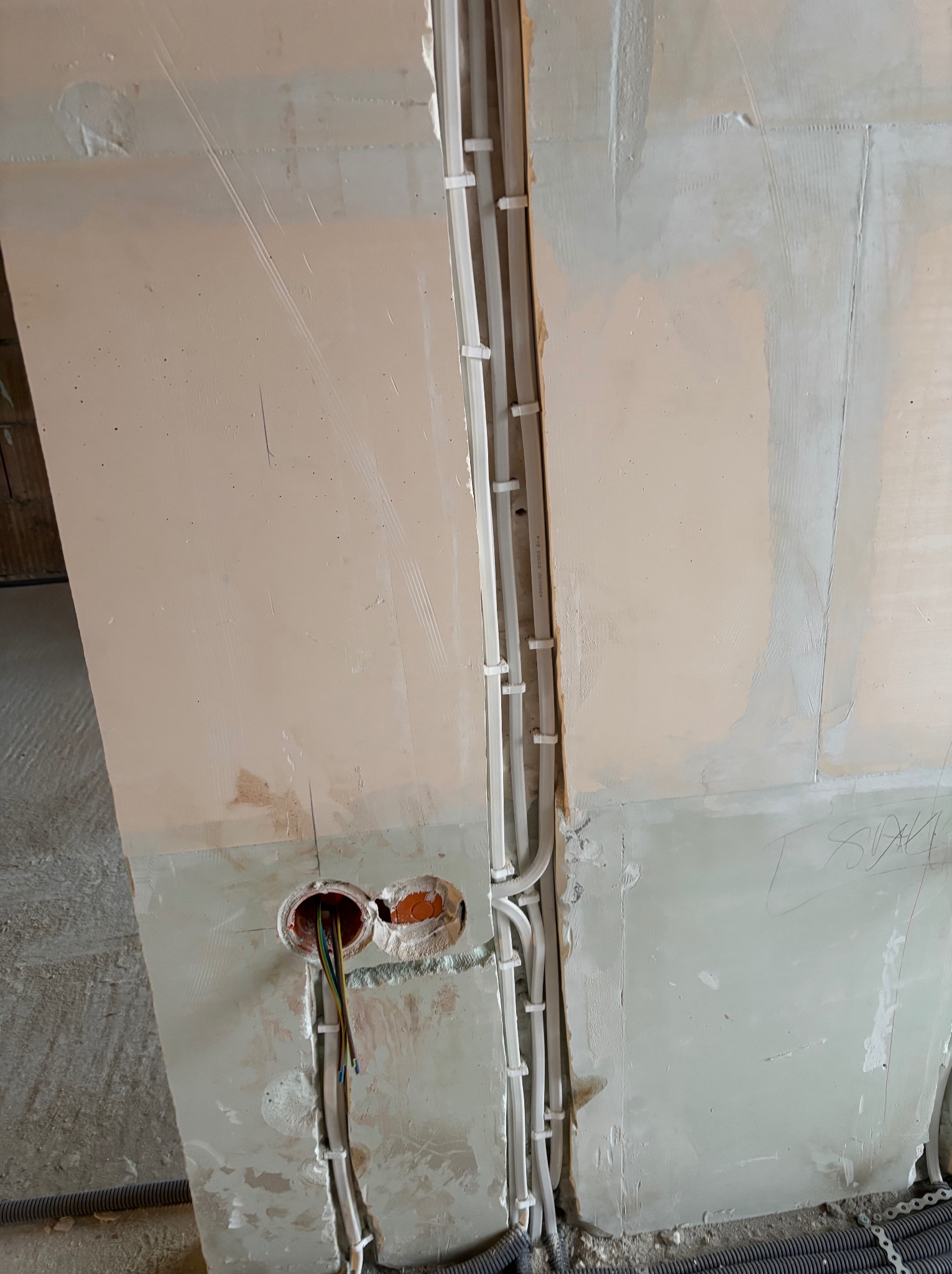 Ściana w trakcie remontu z widocznymi przewodami elektrycznymi w peszlach i puszkami instalacyjnymi. Widoczne kable w kolorach: zielonym, żółtym i niebieskim.