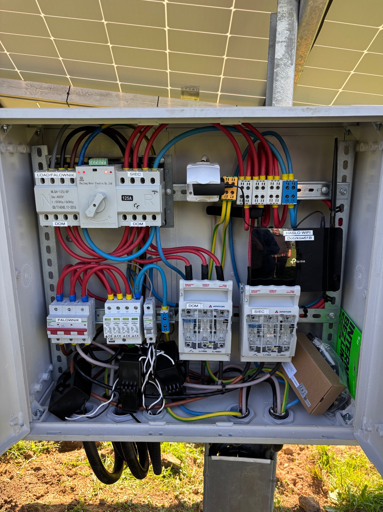 Otwarta skrzynka elektryczna z zabezpieczeniami, przewodami i routerem WiFi. Widoczne bezpieczniki, przełączniki i okablowanie. Na górze fragment paneli słonecznych.
