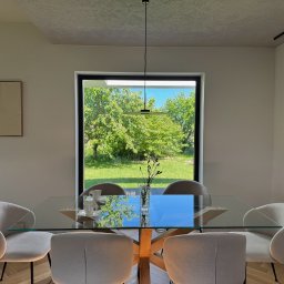 PRACOWNIA PROJEKTOWA KAMIL BUBEL - Jasna jadalnia z parkietem w jodełkę, szklanym stołem i widokiem na ogród przez duże okno w czarnej ramie. Minimalistyczny design, lampa wisząca i dekoracja na ścianie.