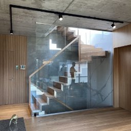PRACOWNIA PROJEKTOWA KAMIL BUBEL - Nowoczesne schody drewniane z szklaną balustradą w minimalistycznym wnętrzu. Widoczne oświetlenie punktowe i drewniana podłoga. Betonowy sufit dodaje surowości.