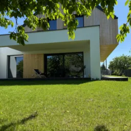 Nowoczesny dom z drewnianą elewacją i dużymi oknami, widok z zadbanego trawnika, fragment korony drzewa w kadrze. Minimalistyczny design, jasne kolory.