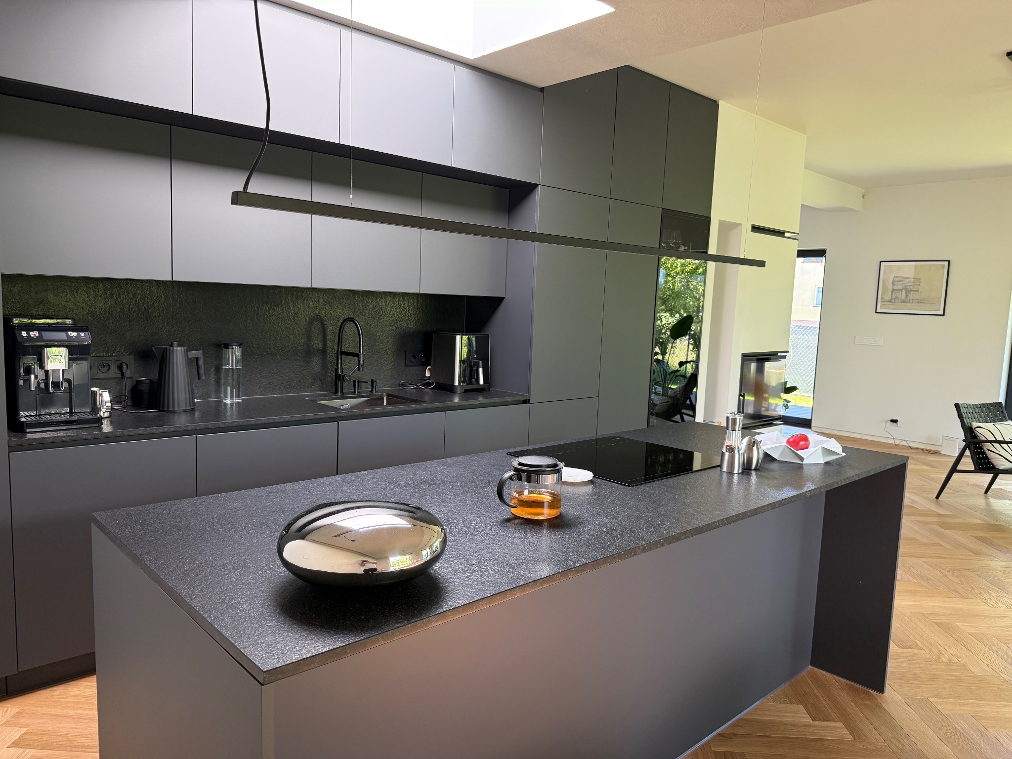 Stylowa, szara kuchnia z wyspą i nowoczesnym oświetleniem. Na blacie czajnik z herbatą i dekoracyjna misa. Parkiet w jodełkę na podłodze. Widok na salon.