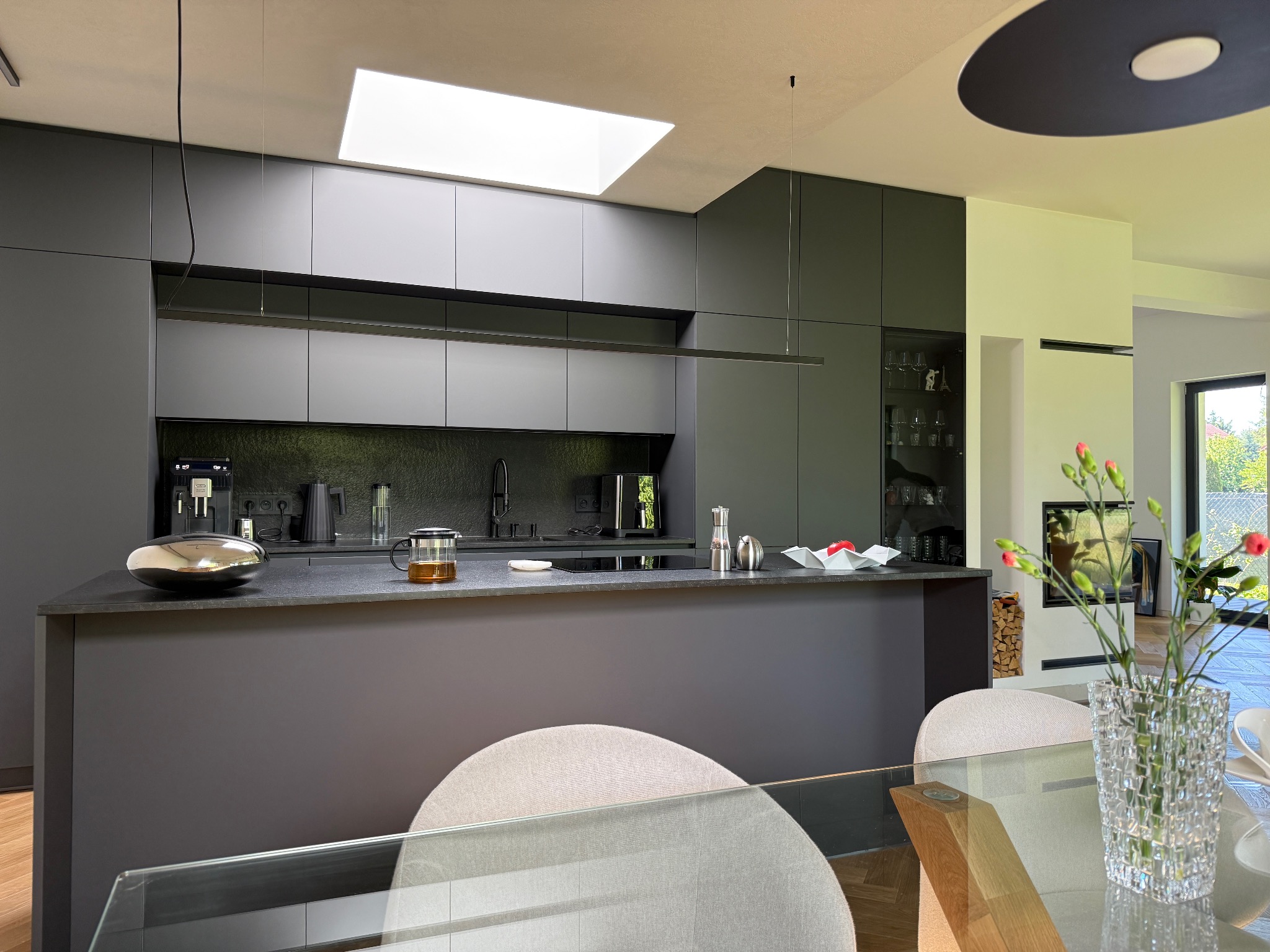 Nowoczesna, szara kuchnia z wyspą i oknem dachowym. Minimalistyczny design z akcentami drewna i szkła. Widoczny fragment jadalni z kwiatami.
