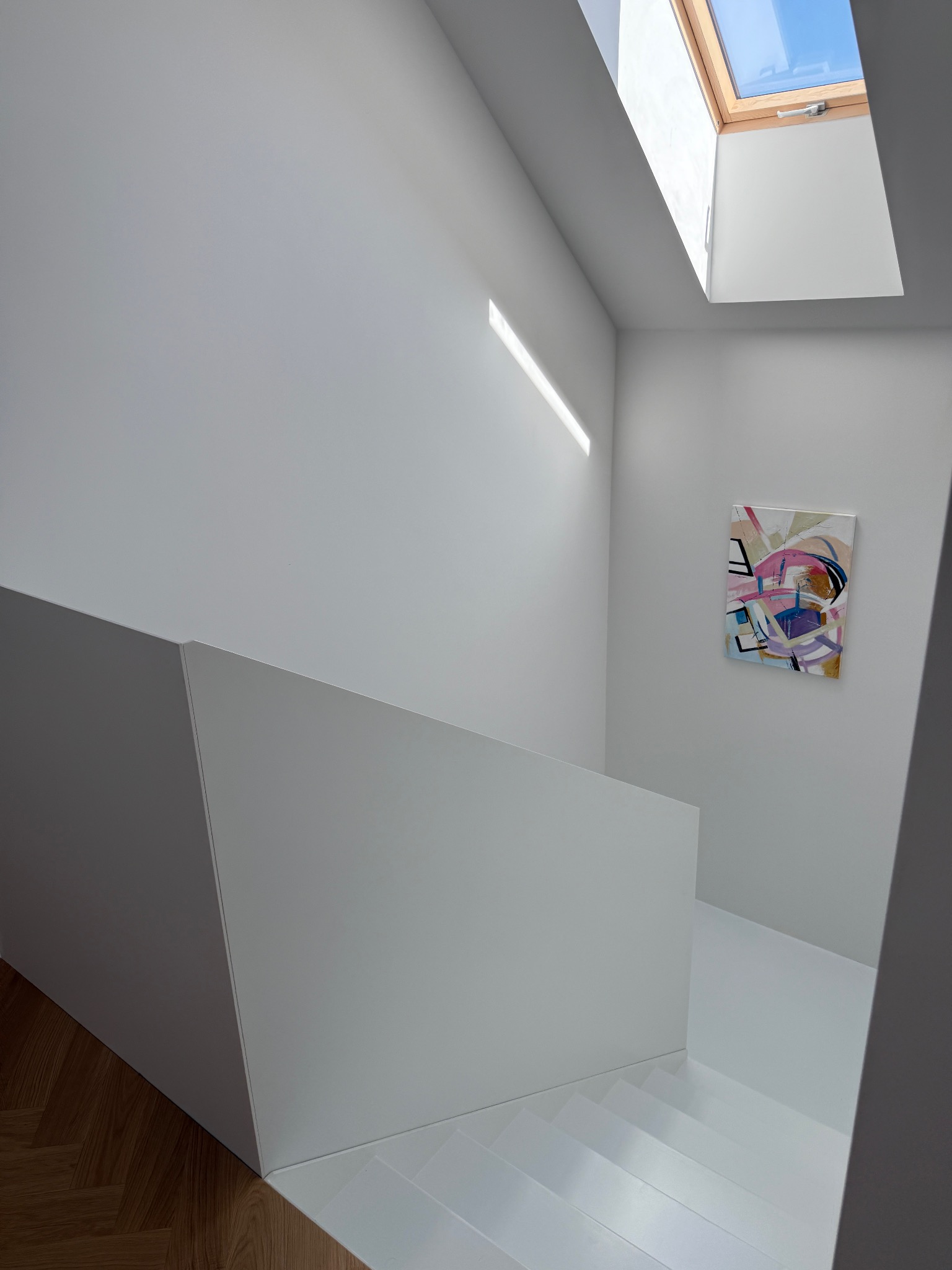 Minimalistyczne wnętrze: białe schody z drewnianym akcentem, abstrakcyjny obraz na ścianie, doświetlone oknem dachowym. Nowoczesny design.