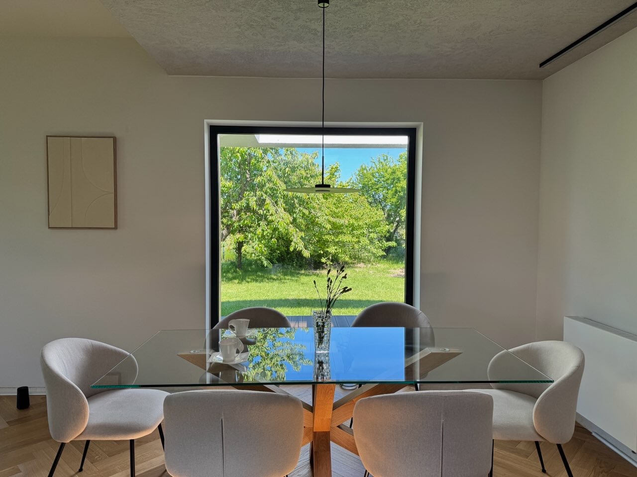 Jasna jadalnia z parkietem w jodełkę, szklanym stołem i widokiem na ogród przez duże okno w czarnej ramie. Minimalistyczny design, lampa wisząca i dekoracja na ścianie.