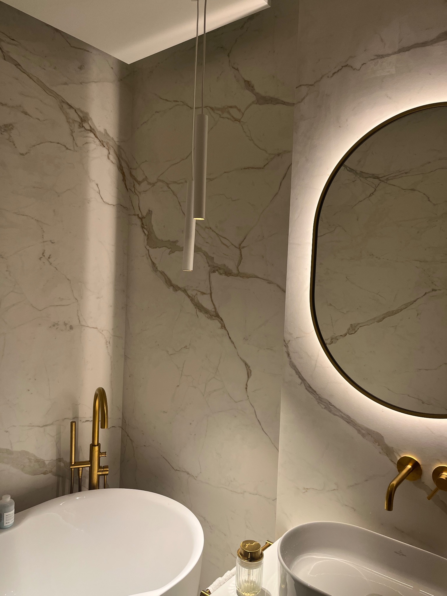 Elegancka łazienka z marmurowymi ścianami, złotą armaturą i okrągłym lustrem z podświetleniem LED. Minimalistyczna lampa wisząca i owalna umywalka.