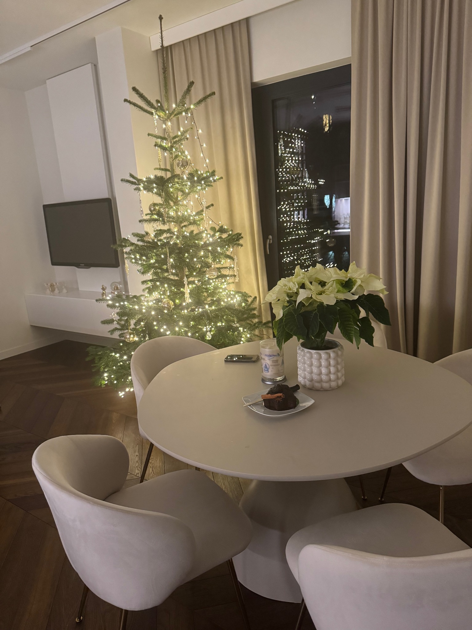 Świąteczny wystrój wnętrza: okrągły stół z ciastem i poinsecją na tle choinki z lampkami i widoku na miasto nocą. Minimalistyczny salon z telewizorem.