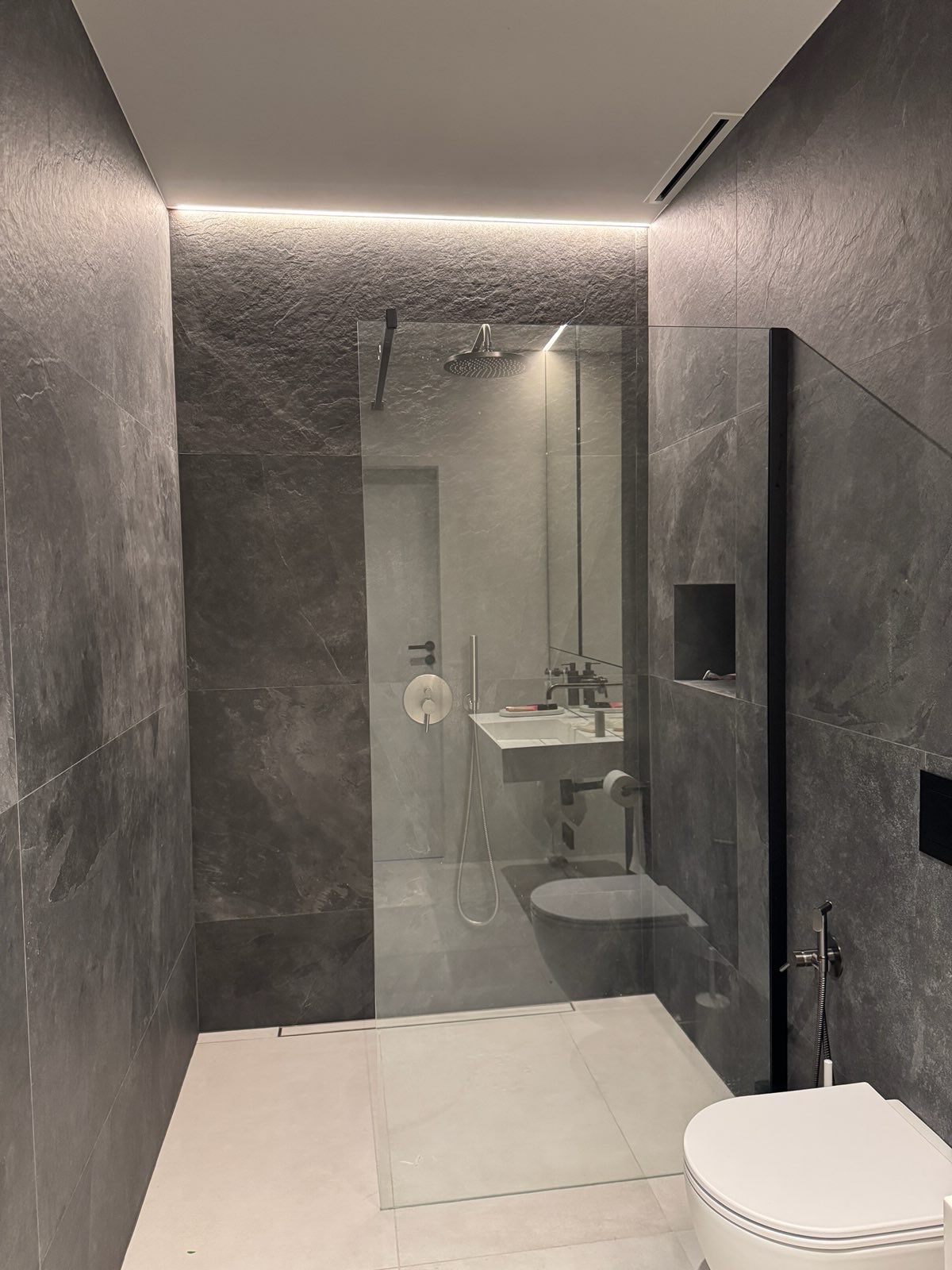 Nowoczesna łazienka z kabiną prysznicową typu walk-in, szare płytki imitujące kamień, podświetlenie LED, biała ceramika i minimalistyczna armatura.