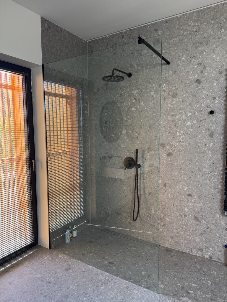 Nowoczesna kabina prysznicowa walk-in z szarymi płytkami lastryko, czarną armaturą i odpływem liniowym. Okno z żaluzjami w tle.