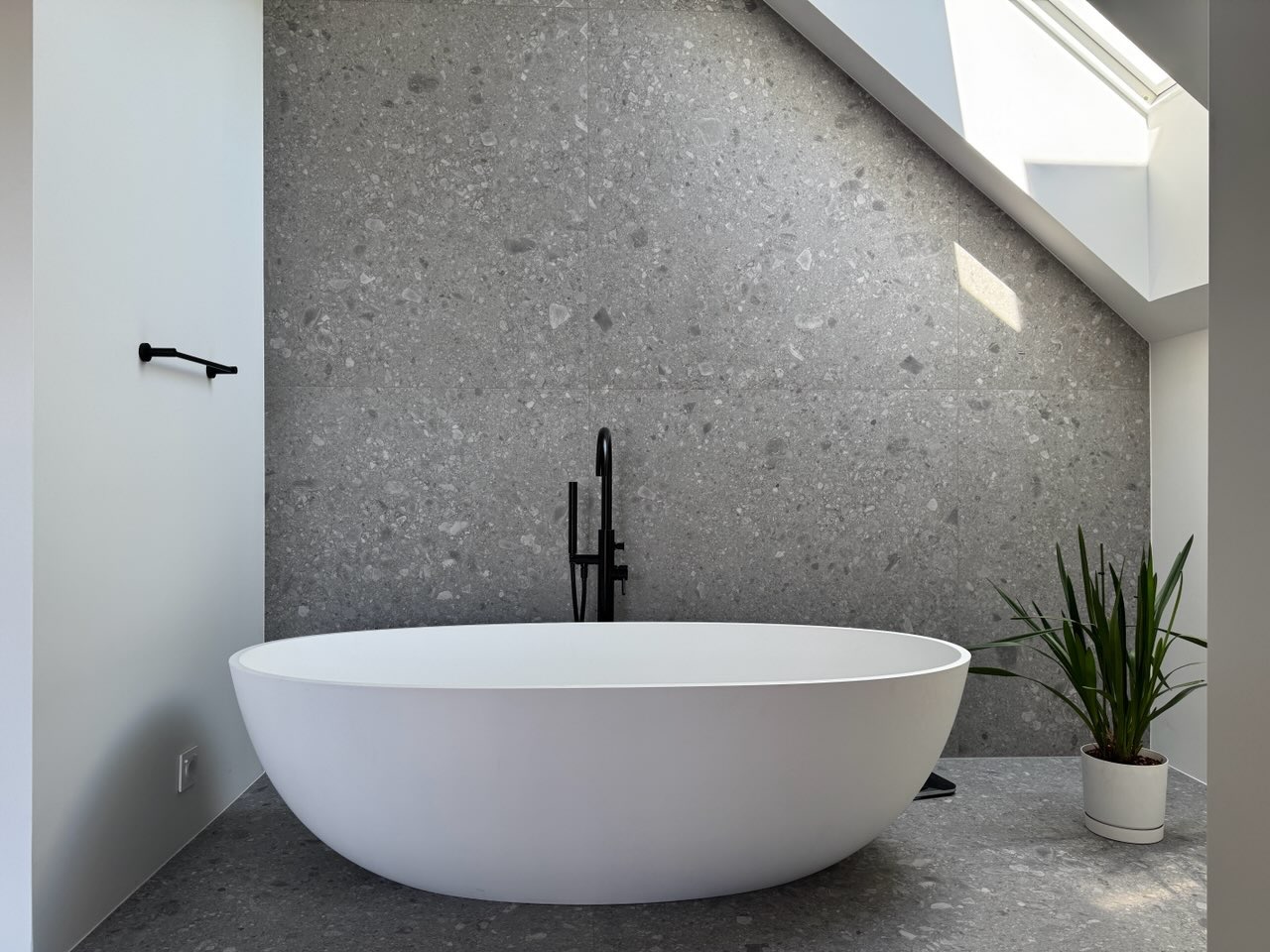 Nowoczesna łazienka z wolnostojącą wanną i szarą mozaiką w Warszawie. Minimalistyczny design, czarna armatura, roślina doniczkowa i okno dachowe.