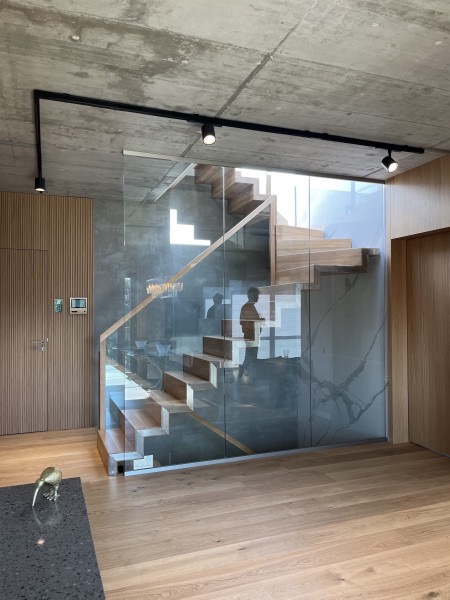 Nowoczesne schody drewniane z szklaną balustradą w minimalistycznym wnętrzu. Widoczne oświetlenie punktowe i drewniana podłoga. Betonowy sufit dodaje surowości.