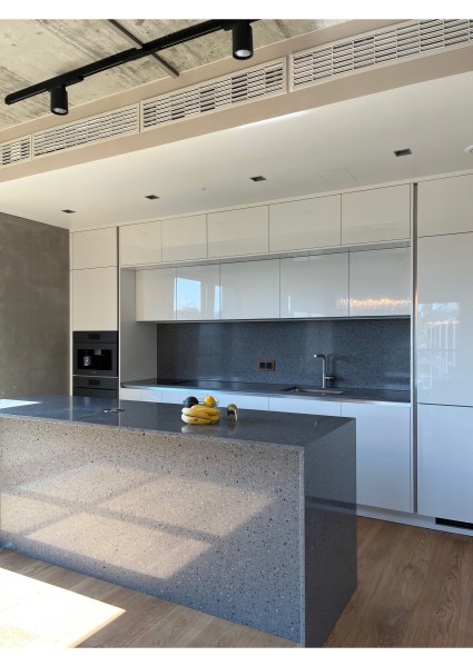 Nowoczesna kuchnia z wyspą: białe szafki z połyskiem, szary blat i backsplash z terazzo, oświetlenie szynowe, owoce na wyspie. Minimalistyczny design.