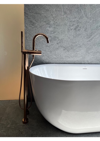 Nowoczesna, wolnostojąca wanna z baterią w kolorze różowego złota na tle szarej, kamiennej ściany. Minimalistyczny design łazienki.