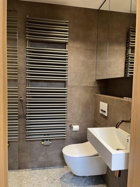 Nowoczesna łazienka z grzejnikiem drabinkowym, wiszącą toaletą i minimalistyczną umywalką na tle brązowych płytek. Widoczny fragment lustra i podłogi z lastryko.