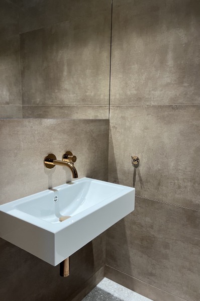 Nowoczesna, biała umywalka z złotą armaturą na tle ściany wyłożonej szarymi płytkami imitującymi beton w łazience. Minimalistyczny design.