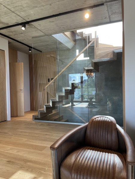 Nowoczesne wnętrze z drewnianymi schodami za szklaną ścianą. W tle widoczne drzwi i krzesło w stylu vintage. Oświetlenie sufitowe punktowe, betonowy sufit.