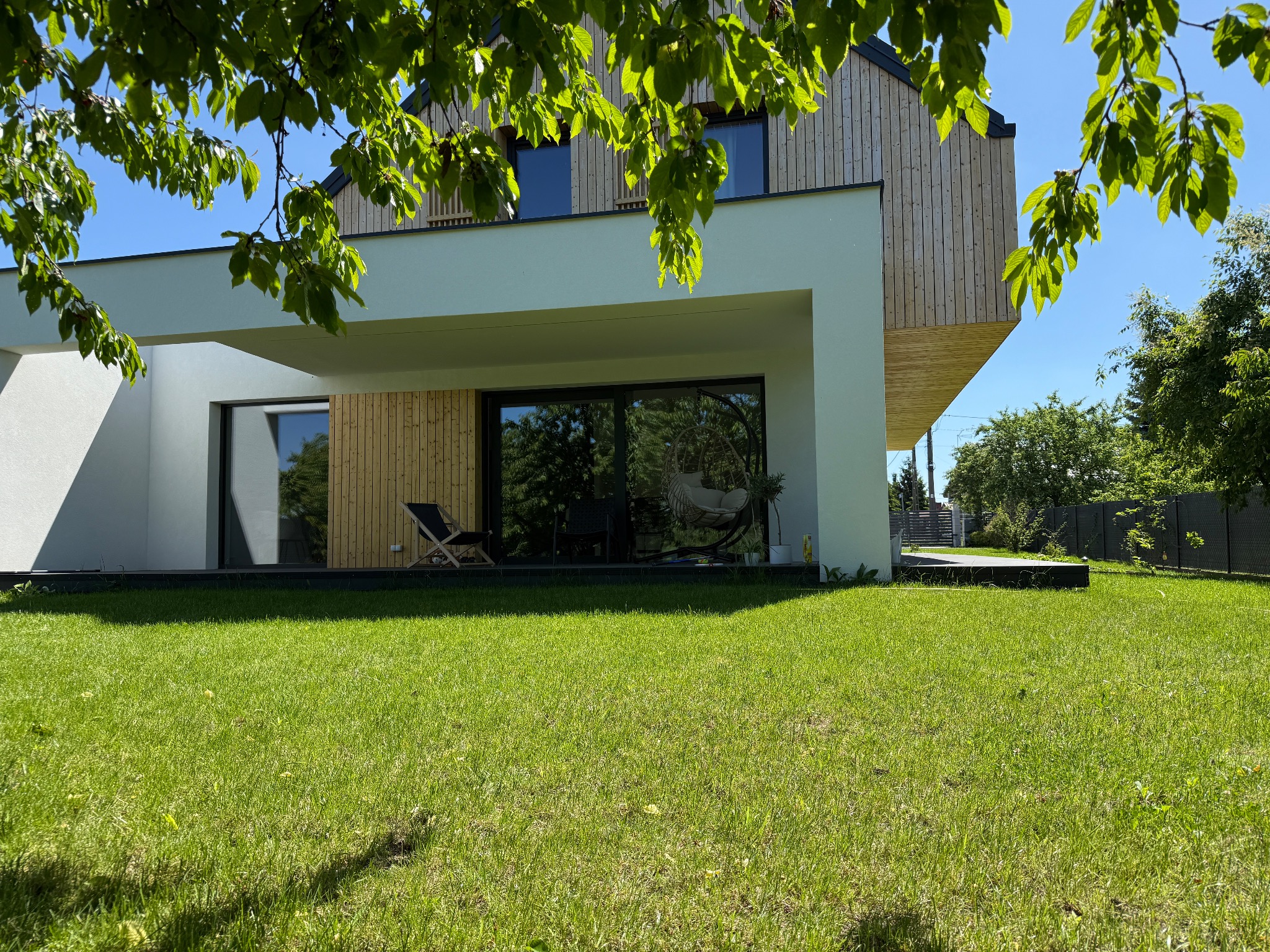 Nowoczesny dom z drewnianą elewacją i dużymi oknami, widok z zadbanego trawnika, fragment korony drzewa w kadrze. Minimalistyczny design, jasne kolory.