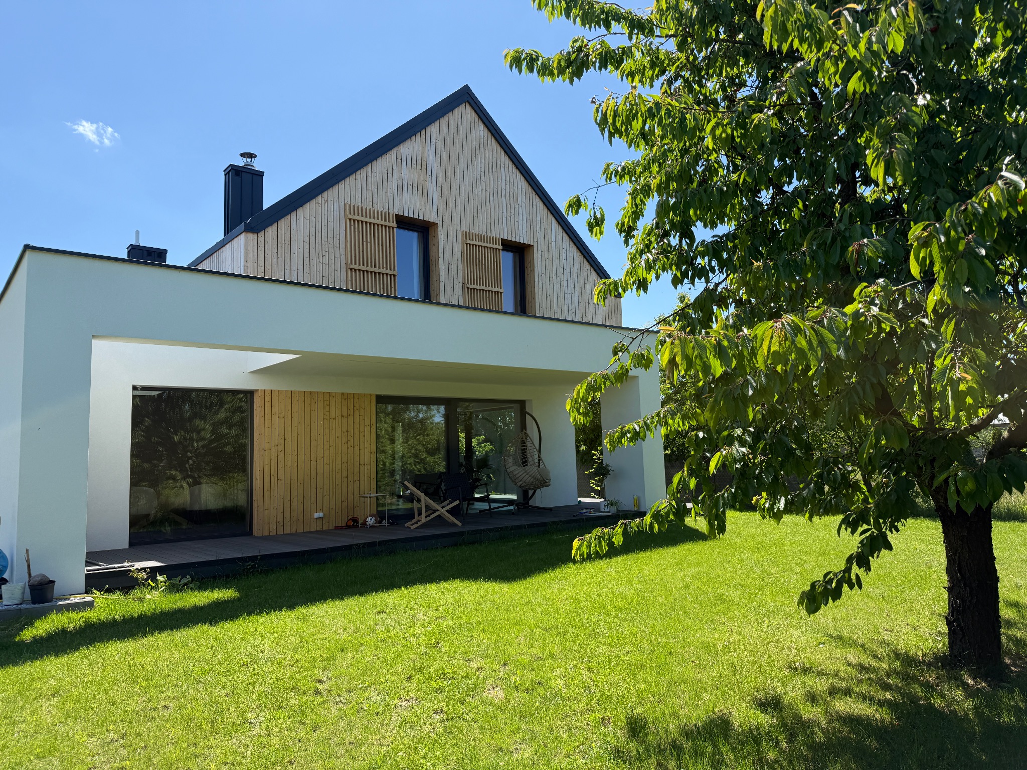 Nowoczesny dom z drewnianą elewacją i białym, minimalistycznym gankiem, otoczony zielonym trawnikiem i drzewem. Widok z zewnątrz w słoneczny dzień.