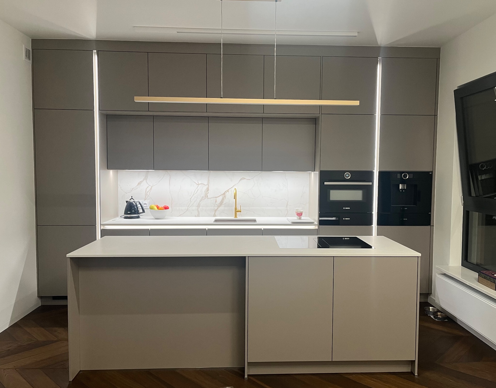Nowoczesna kuchnia w odcieniach szarości z wyspą, oświetleniem LED i marmurowym backsplashiem. Uwagę przykuwa złota bateria i minimalistyczna lampa wisząca.