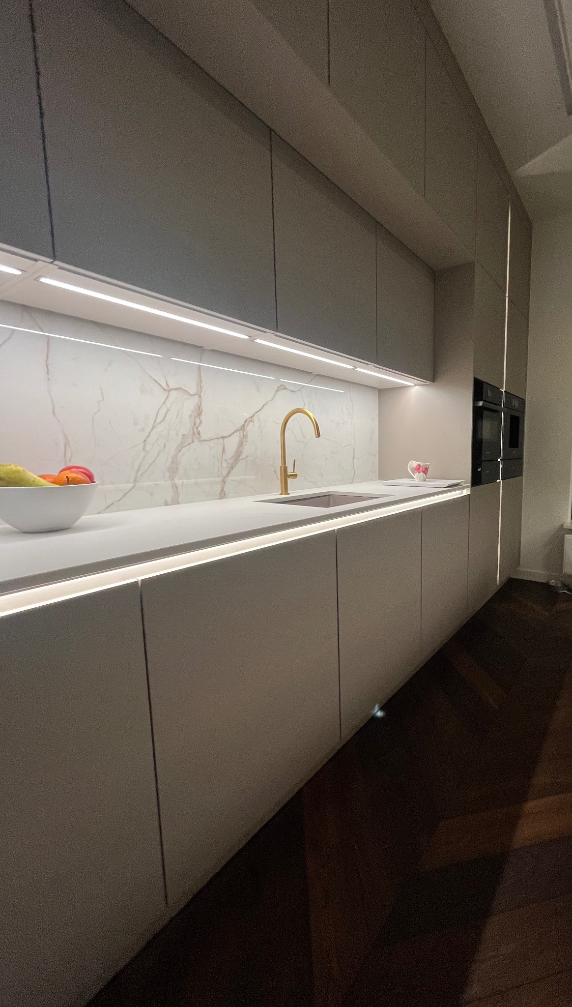 Nowoczesna kuchnia z szarymi szafkami, marmurowym backsplashiem i złotą armaturą. Oświetlenie LED podkreśla blat i przestrzeń roboczą. Parkiet na podłodze.